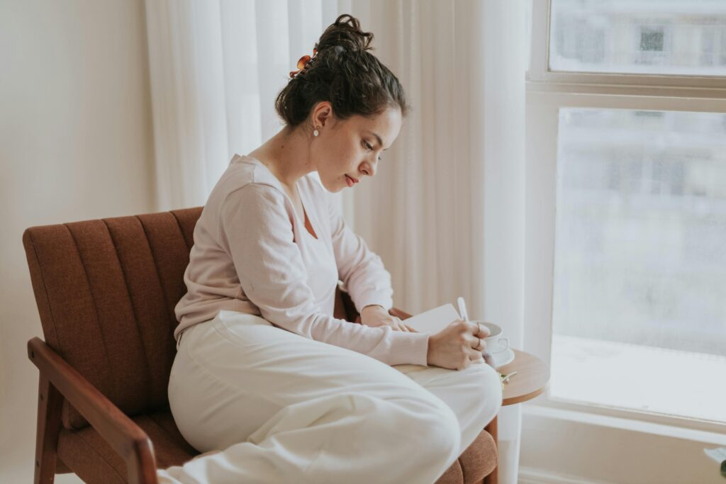 A woman journaling beside a sunlit window, reflecting on personal growth.